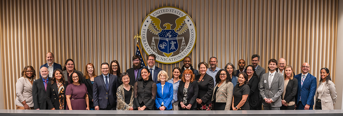 2024 Consumer Advisory Committee members in the FCC commission room - click to enlarge